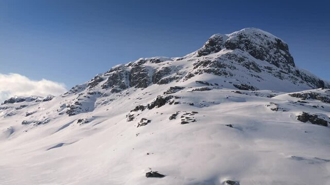 Aerial approaching shot of snowy Bitihorn mountain in Norway at sunny day. Snow covered mountain peak against blue sky. BOttom Up shot.