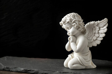 Fototapeta premium A marble angel statue sitting in prayer on a slate surface with a dark background. Perfect for spiritual, memorial, or romantic designs with space for text.