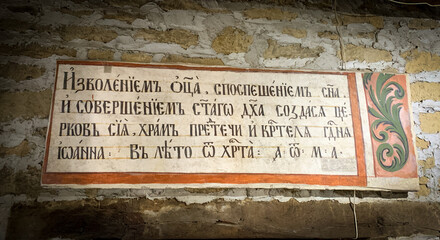 A historical inscription in Cyrillic script is displayed on an old stone wall, bordered by a decorative frame with floral patterns on the right side.