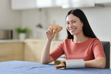 Happy asian woman eating cookies looking at you in the kitchen
