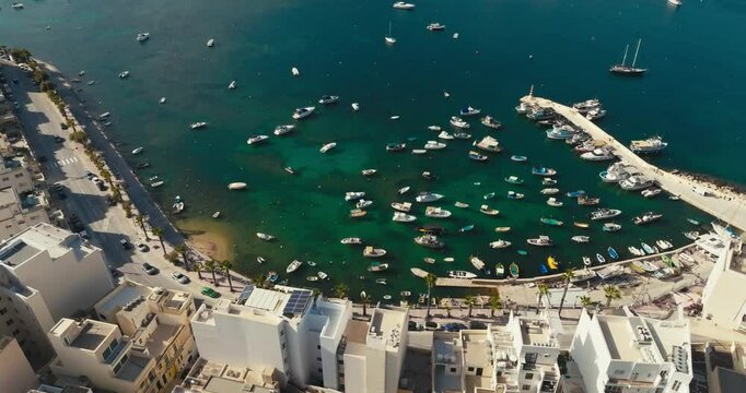 Aerial view of the marina in Bugibba, Malta. Yachts and fishing boats are moored near the pier on the promenade of the Mediterranean tourist town. High quality 4k footage