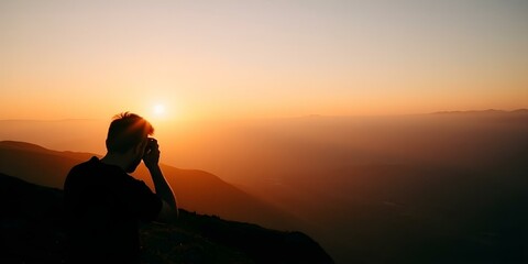 Obraz premium A silhouette of a person sitting on a mountain peak at sunset, with the sun peeking through the hazy sky and mountains in the background