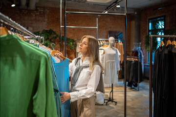 Interested woman buyer carefully examines clothing for new season, picking blouses in shop. Pleased middle aged female customer attentive looking outfit before purchase, make choice standing in store.
