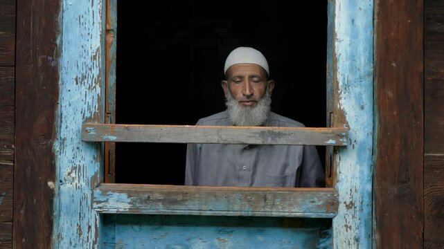 An aged or elderly Muslim Indian Kashmiri man in ethnic attire sitting indoors, looking outside from the frame of the window of an old village wooden house in a Himalayan mountainous region of Kashmir