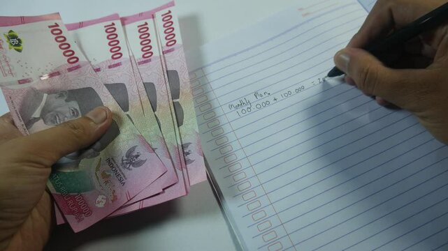Hand writing financial notes while holding Indonesian rupiah banknotes. Concept of personal finance management, budgeting, accounting, and money planning with a realistic close-up view.