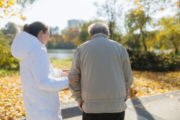 Obraz premium Elderly man and nurse in autumn park, back view of outdoor companionship