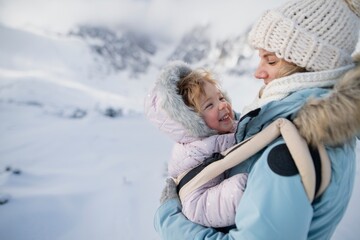 Fototapeta premium Mother standing in the middle of snowy landscape with daughter in baby carrier. Mom and girl enjoying winter holiday in the mountains.