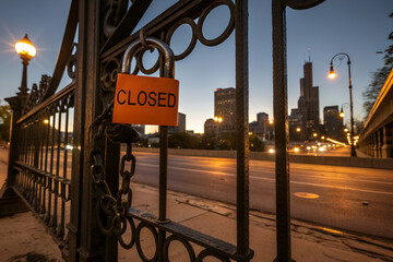 A padlocked gate with a "CLOSED" sign, set against a city skyline at dusk, evoking a sense of abandonment and stillness in an urban environment.