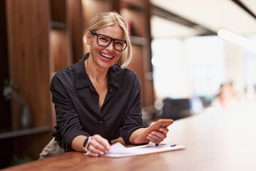 A confident, smiling professional woman sits at her desk, surrounded by a phone and documents