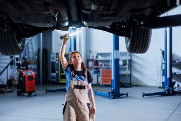 Skilled repairman working on suspended car in garage, checking tires during routine maintenance. Adept auto repair shop specialist underneath vehicle, inspecting it using work light