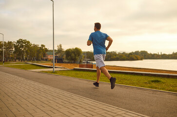 A sporty guy runs at dusk through the city near the lake, back view. A male sprinter trains alone....