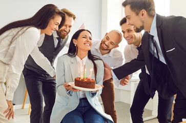 Team of business people celebrating a colleague's birthday at work. Group of coworkers and friends giving a cake with candles to a happy, smiling young woman in a party cap