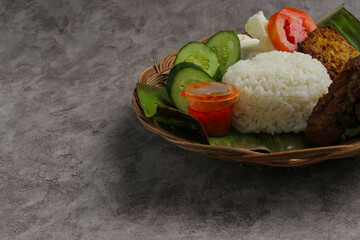 Delicious plate of Indonesian food featuring ayam bakar (roasted chicken), served with a mound of white rice, crispy cracker, tempeh, and fresh vegetables like cucumber and tomato on a woven basket