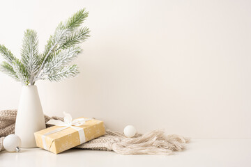 Minimalist side view Scandinavian style Christmas scene featuring a gift wrapped with ribbon, frosted pine branches in a vase, and cozy knit blanket