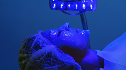 Young woman having blue LED light facial therapy treatment in beauty salon. Beautician wearing face...