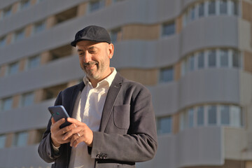 Portrait of elegant smiling businessman in cap looking at smart phone in city with buildings on background