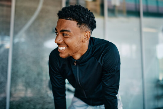 Fit young man in sports clothing looking sideways and smiling while sitting on the floor of a gym during a break from a workout session
