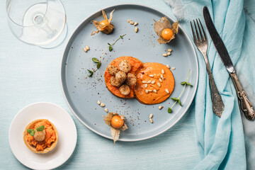 A gourmet scallops lunch with carrot puree in Romesco sauce served on a blue plate with fesalis, a glass of white wine and cutlery.