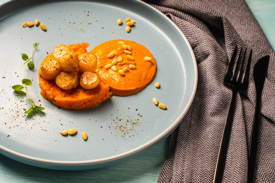 Scallops with carrot puree with Romesco sauce, fesalis and pine nuts on a blue plate on a light background with a napkin and cutlery. Diet food, vegetarian dish