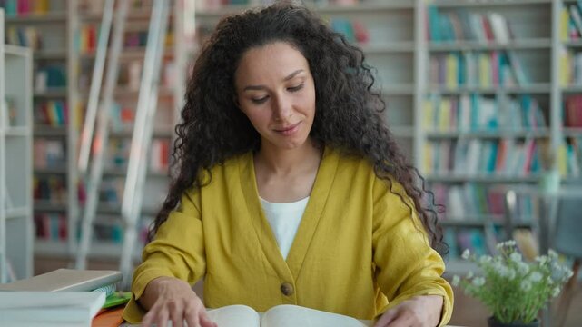 Hispanic Arab Indian woman reading book reader pages library looking at camera smile girl student teenager textbook female knowledge literature academic bookworm bookshelves educated study bookstore