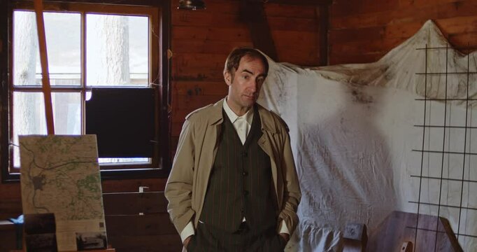 A spy dressed in a classic trenchcoat gives a briefing during 1940s World War 2. A map with information and details is seen in the background of the hide out