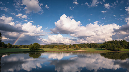 Fototapeta premium A serene lake reflects fluffy clouds and a clear blue sky, with a distant rainbow and lush green trees lining the shore.