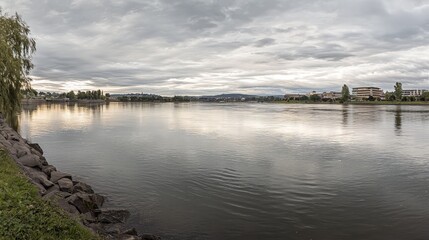 Obraz premium Panoramic view of calm river reflecting cloudy sky, cityscape in background.