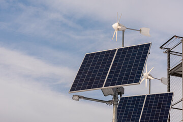 Na zdjęciu są panele słoneczne i małe turbiny wiatrowe, które produkują energię z odnawialnych źródeł. Widać niebo z chmurami i fragment metalowej kratownicy obok urządzeń © Marcin