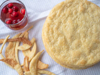 Top view over a pile of baked thin round buttery layers, dough scraps on a white waffle towel ready to make a shortcrust puff pastry Napoleon cake. A glass of red berry drink nearby.