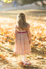 back of little girl with long blonde hair with fancy floral dress standing outside in the foliage on a fall day 