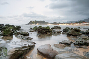 A serene coastal view with green moss-covered rocks in the foreground, calm ocean waves, and a distant hill with lights and buildings. The cloudy sky creates a peaceful and scenic setting.