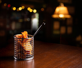 fried sweet potato chips in stainless steel fryer basket isolated on wooden table in blurred restaurant corner at night