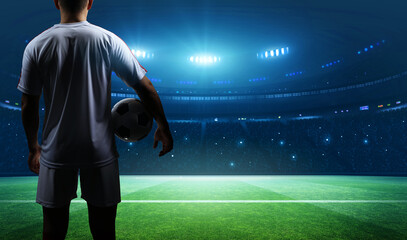 Soccer player standing in a stadium, facing away and holding a ball.	