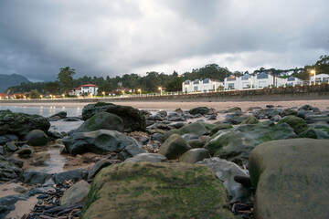 Coastal view with moss-covered rocks, sandy beach, and white buildings along a promenade. Cloudy sky and soft lighting create a serene and inviting atmosphere, perfect for travel and tourism themes.