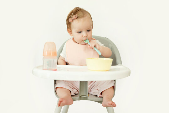 Cute little baby girl sitting on high chair and taking spoon to mouth, feeding herself with porridge or mush fruit puree