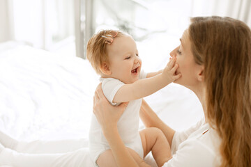 Young mother playing with her baby girl at home, sitting in the bedroom, kid touching mom's face and smiling, side view, free space