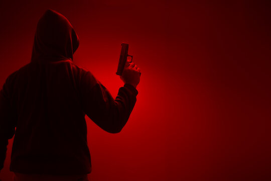 An unidentified man in a black hood and black mask stands holding a gun in various poses. Light is shining from above. Looks scary. Isolated over film studio background. Armed person.