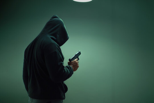 An unidentified man in a black hood and black mask stands holding a gun in various poses. Light is shining from above. Looks scary. Isolated over film studio background. Armed person.