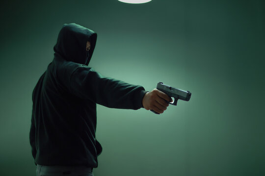 An unidentified man in a black hood and black mask stands holding a gun in various poses. Light is shining from above. Looks scary. Isolated over film studio background. Armed person.