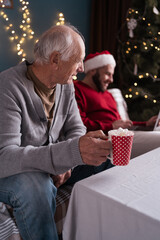 Busy son working online on christmas visiting old father sitting on sofa. Elderly man feels lonely...