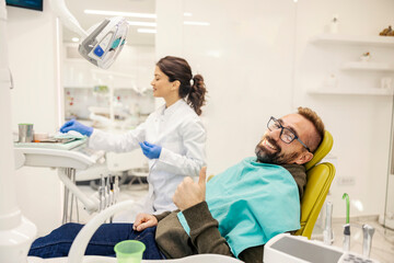 Smiling man sitting in char at dentist office and giving thumbs up while dentist in a blurry...