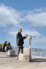 A man stands next to a coin-operated telescope on a scenic lookout, wearing a black leather jacket, camouflage pants, and reflective sunglasses. Visible individuals tourism, adventure, and leisure