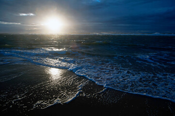 A glowing sunrise over the ocean, with sunlight reflecting on gentle waves and dark clouds overhead, creating a tranquil and dramatic coastal scene.