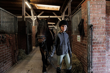 Young little teenager kid girl leading chestnut horse through stable ranch yard. Horse riding...