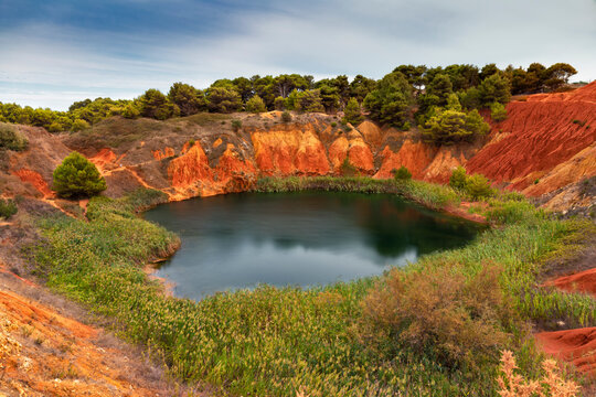 View of bauxite cave with serene lake, red rocks, and lush vegetation, Spinazzola, Italy.