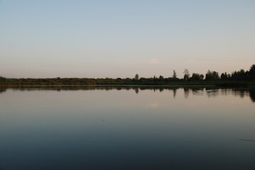 Naklejka premium Windless evening on the lake. Summer evening clear light blue sky without clouds. Smooth flat surface of water in forest lake. Sky and plants growing on opposite shore of lake are reflected from water