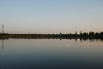 Naklejka premium Windless evening on the lake. Summer evening clear light blue sky without clouds. Smooth flat surface of water in forest lake. Sky and plants growing on opposite shore of lake are reflected from water
