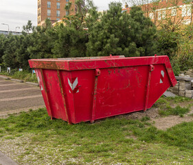 red steel open industrial waste container is placed outside on the grass next to a forest of pine...