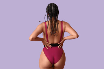 Young African-American woman in stylish purple swimsuit on lilac background, back view