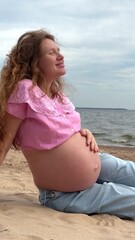 beautiful pregnant woman with big belly and long hair sits on sand by sea, water and enjoys weather